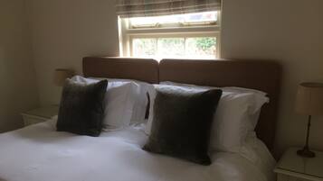 A neatly made bed with white linens and two dark pillows, a bedside table with a lamp, and a window with blinds.