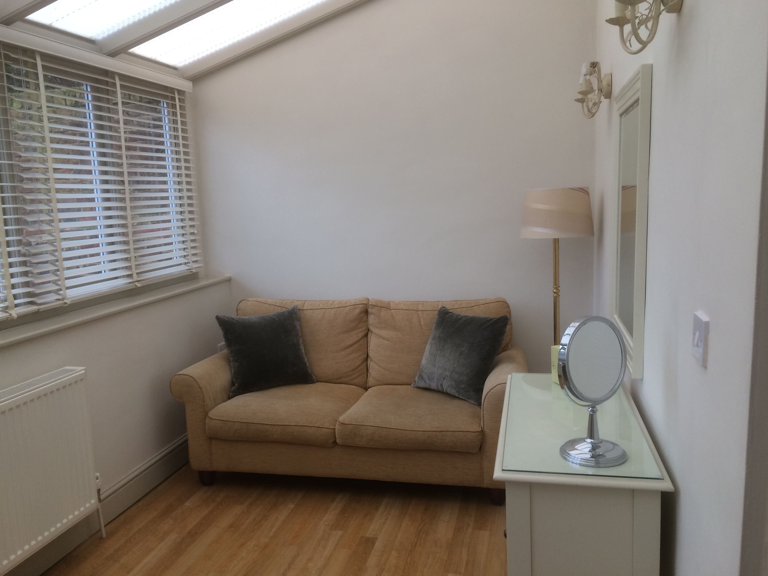 A room with a beige sofa, two dark cushions, a small white side table with a mirror, and a floor lamp.