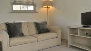 A living room with a beige sofa, two dark cushions, a lamp, a television on a stand, and a window with a striped valance. - The Barn At Woodview (Tonbridge)