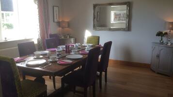 A dining room with a wooden table set for four, surrounded by chairs. A mirror hangs on the wall above the table, and a cabinet is visible to the right.