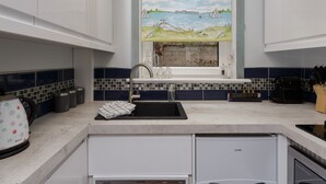 A kitchen with white cabinets, a tiled backsplash, a stainless steel sink, and a window with a painting of a landscape. - Albany Garden (Portsmouth)
