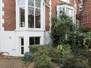 A corner of a building with a white facade, a bay window, and a small garden area with various plants and a bird feeder.