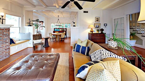 A spacious living room with a leather ottoman, a beige sofa with patterned cushions, a dining area with a wooden table, and a ceiling fan. - Walk to Beach & Pleasure Pier - Lush Outdoor Entertaining (Galveston)