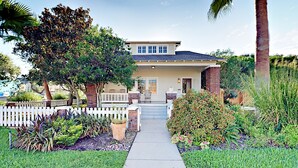 A two-story house with a front porch, a picket fence, and a well-maintained garden with various plants and a palm tree. - Walk to Beach & Pleasure Pier - Lush Outdoor Entertaining (Galveston)