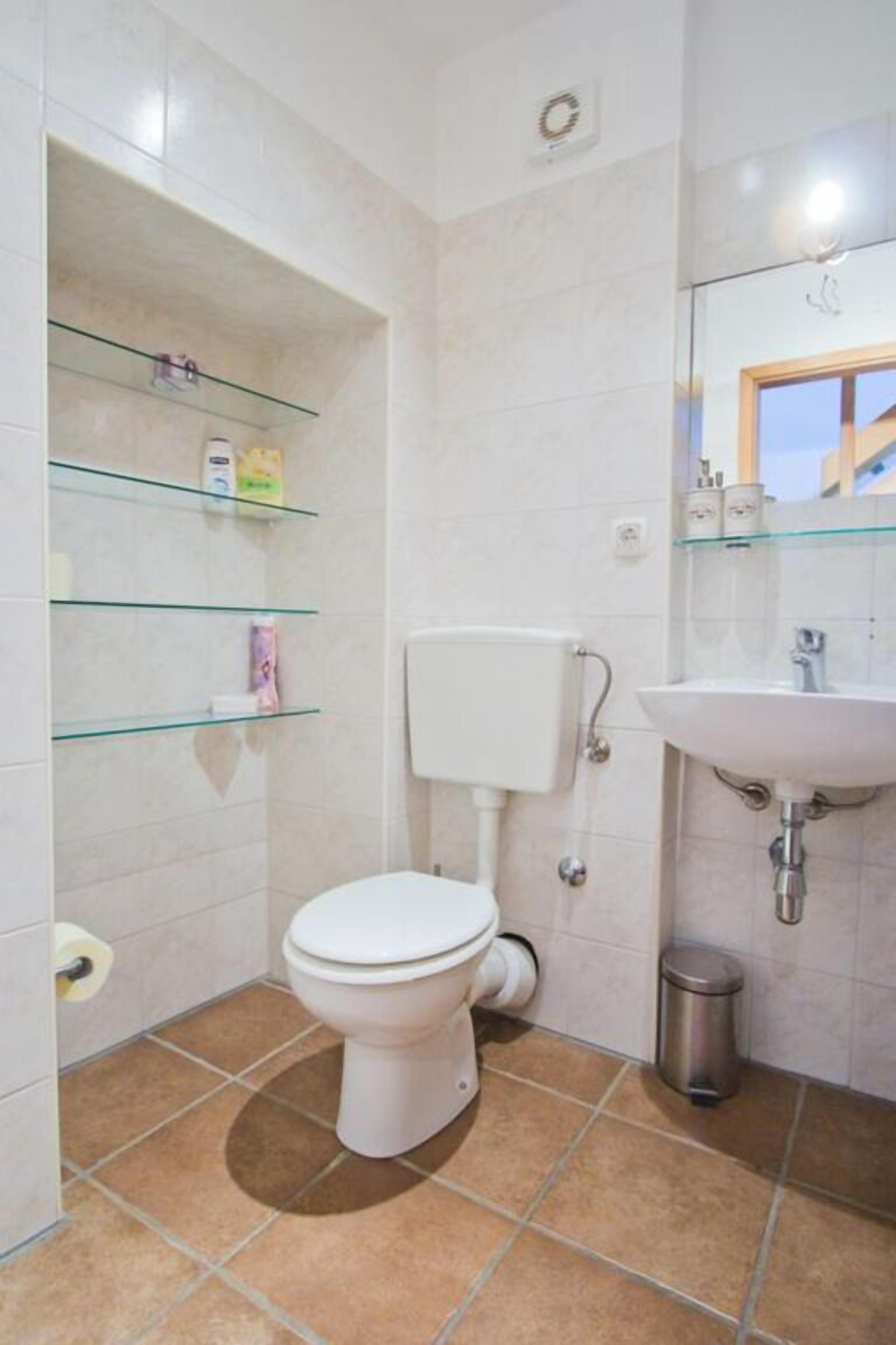 A small bathroom with a toilet, a glass shelf with toiletries, a sink, and a mirror.