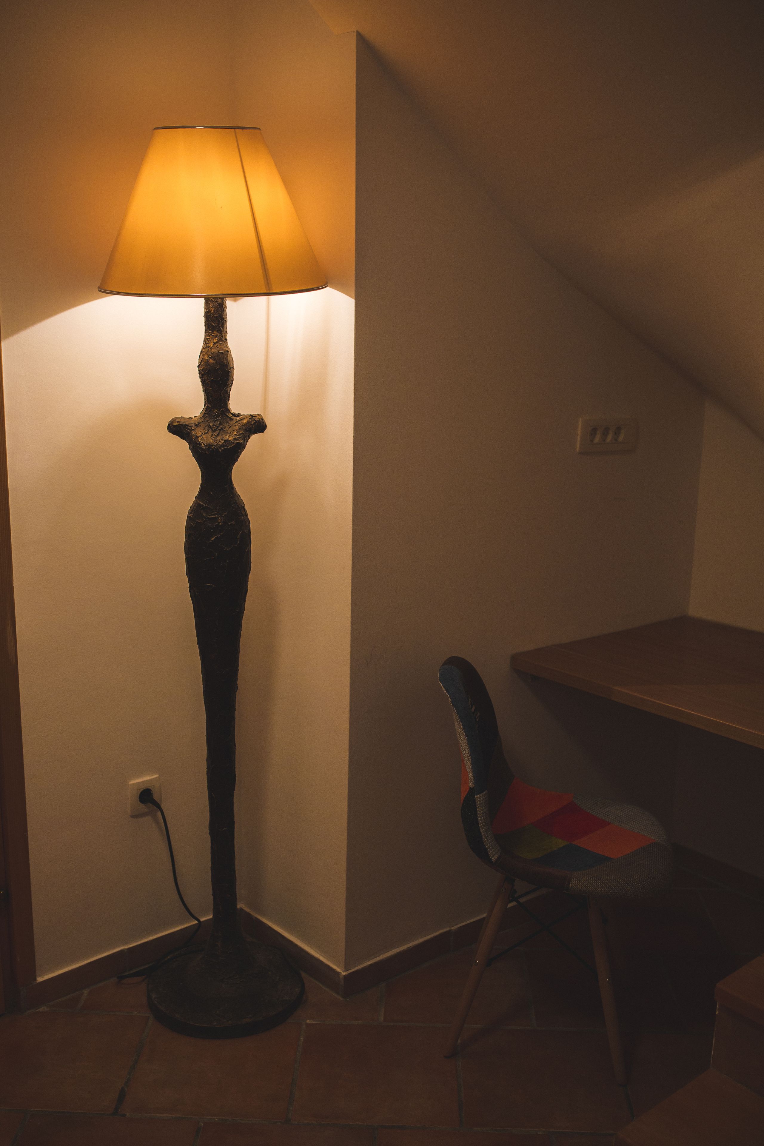 A lamp with a textured base and a conical shade, positioned next to a chair with a colorful cushion.