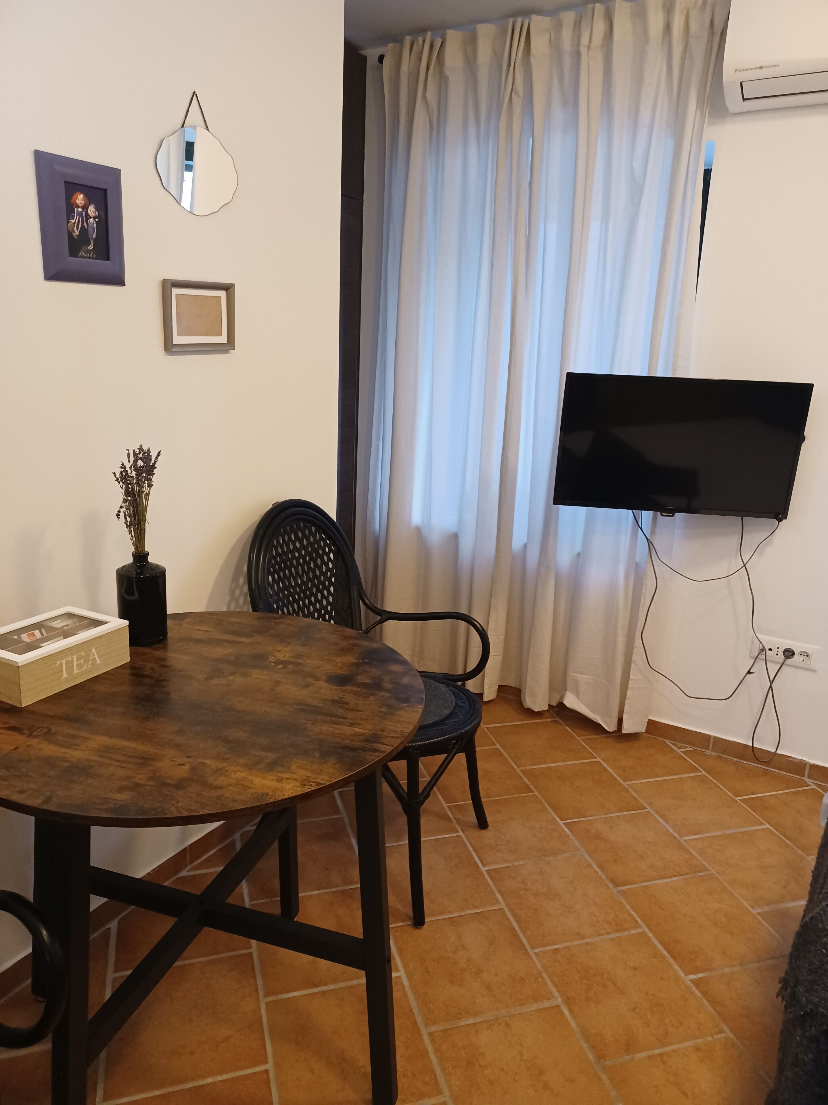 Een woonkamer met een ronde houten tafel, een zwarte stoel, een flatscreen televisie aan de muur, een potplant en een lijst met een foto.