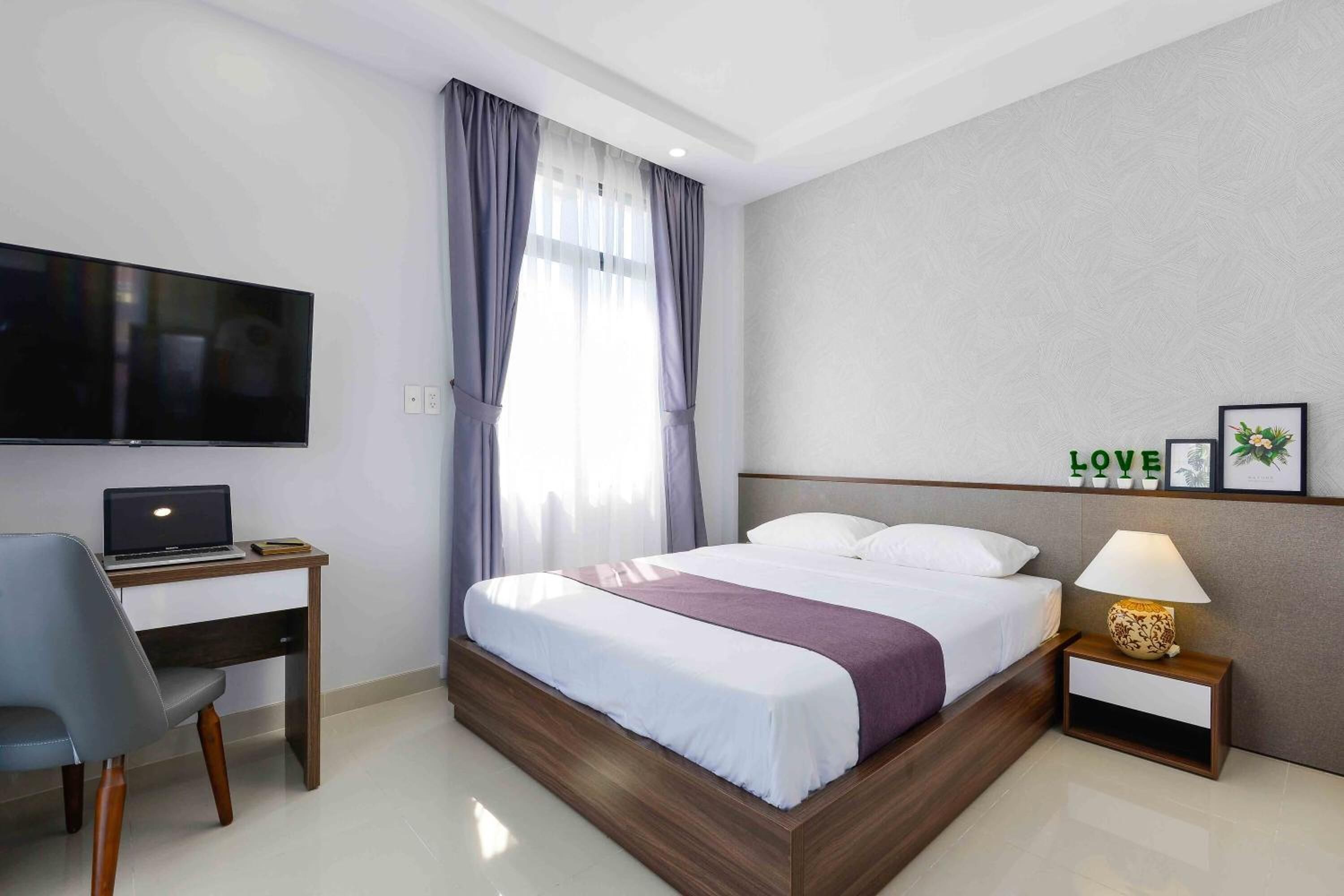 A modern hotel room with a bed, a desk with a laptop, a TV, and a nightstand with a lamp.