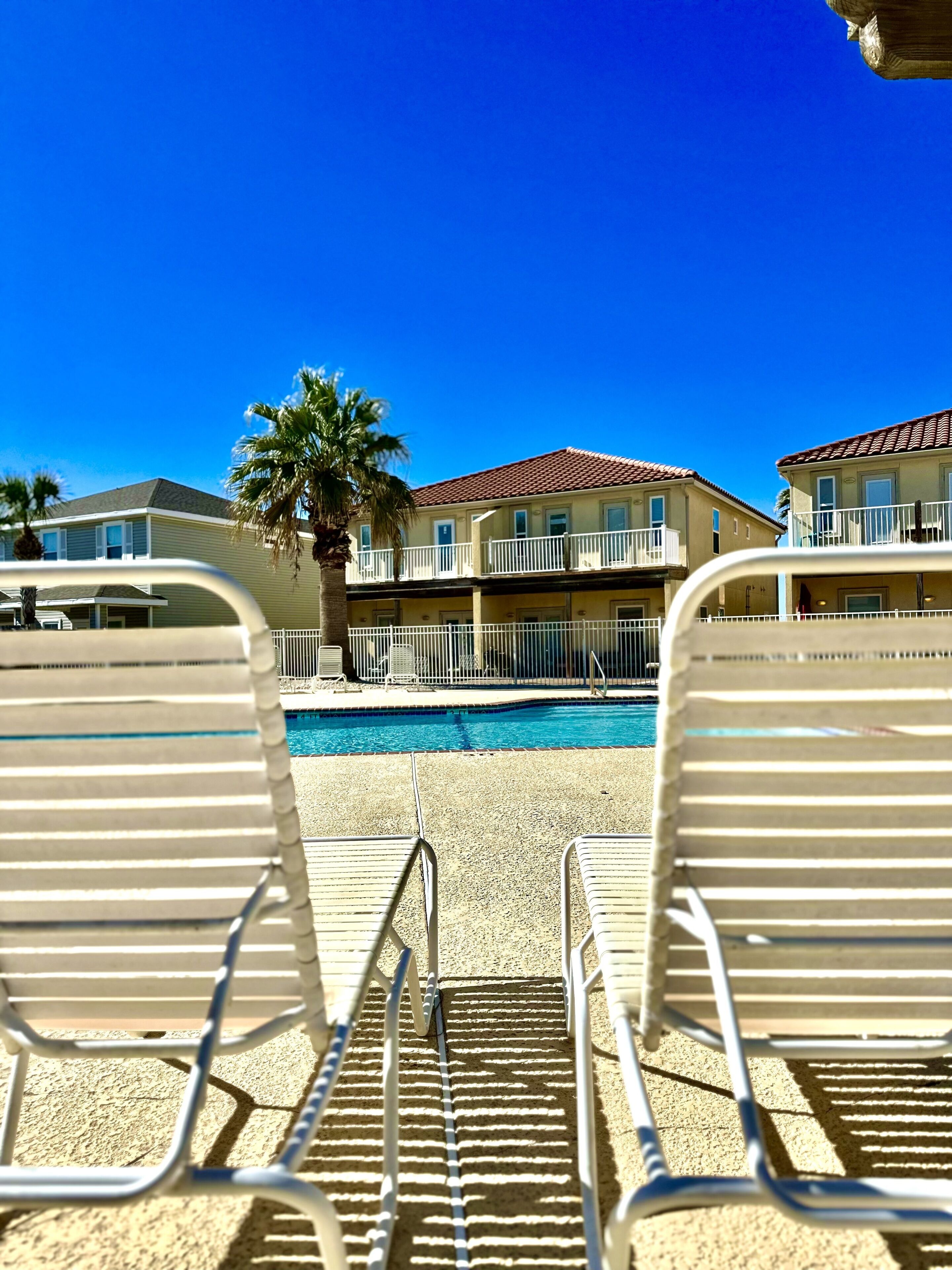 Une vue sur la piscine, avec des chaises longues, un ciel bleu dégagé et un bâtiment doté de balcons.