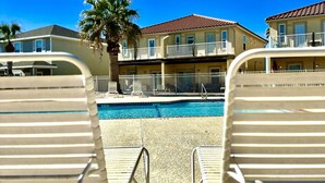 Vista de la piscina con sillas de descanso, cielo azul despejado y un edificio con balcones.