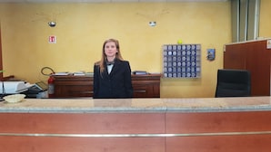 A reception desk with a person standing behind it, a fire extinguisher, a fire exit sign, a bulletin board with numbered cards, and a wooden desk with files and a computer. - RistHotel Airport (Fogliano Redipuglia)