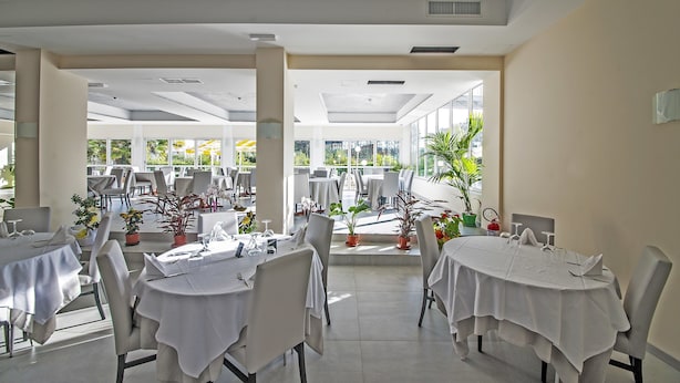 A spacious indoor dining area with tables set for guests, large windows offering views of the outdoors, and potted plants adding greenery. - RistHotel Airport (Fogliano Redipuglia)