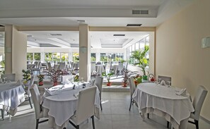 Un vaste espace intérieur aménagé pour les repas, avec des tables dressées pour les convives, de grandes fenêtres offrant une vue sur l’extérieur et des plantes en pot qui apportent une touche de verdure.