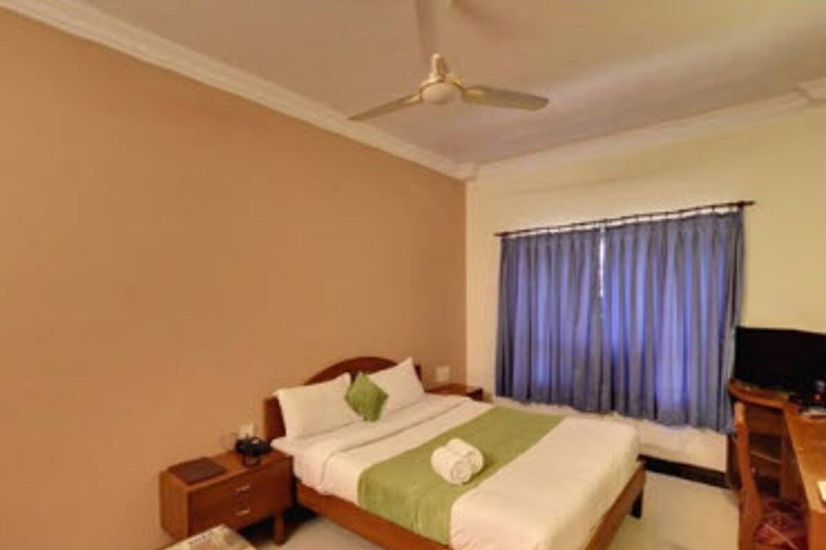 A hotel room with a bed, a nightstand, a desk, a ceiling fan, and a window with curtains.
