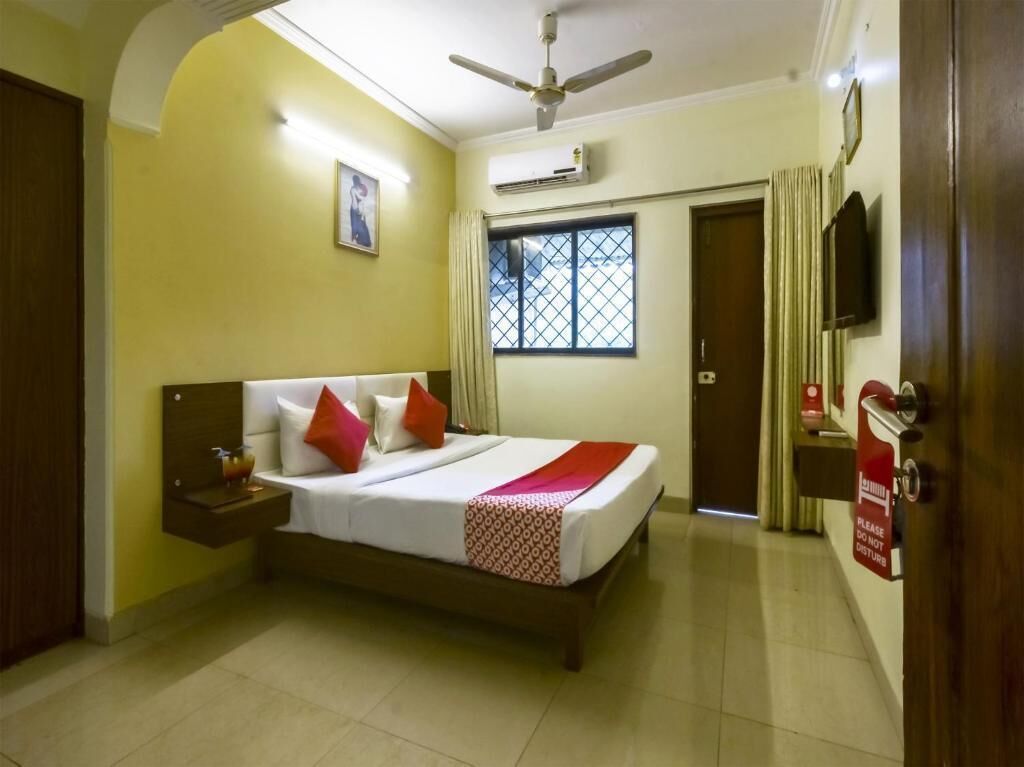 Habitación de hotel con cama, mesita de noche, ventilador de techo, equipo de aire acondicionado, ventana con barrotes, puerta y un cartel rojo de 'No molestar'.
