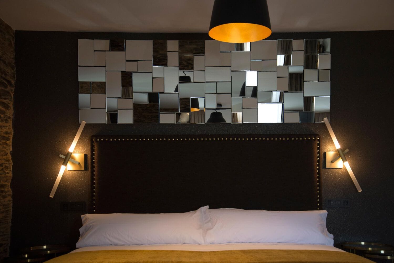 A hotel room with a modern headboard, a large mirror with a geometric pattern, and two bedside lamps.