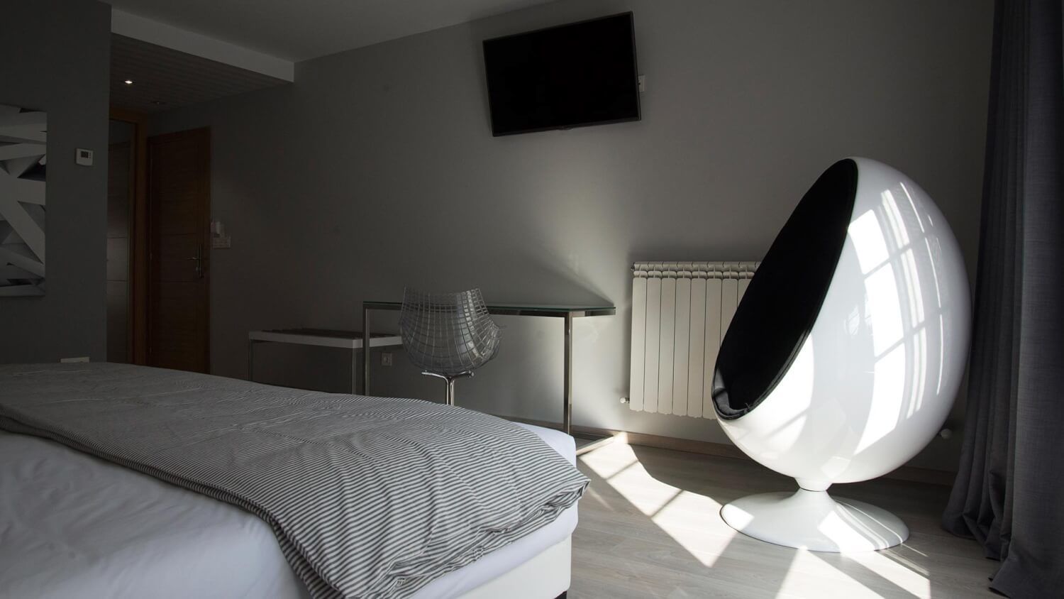 Une chambre moderne avec un lit, une chaise, un fauteuil original en forme d’œuf, un radiateur et un téléviseur fixé au mur.