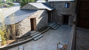 A stone building with a sloped roof, a small window, and a door. There are steps leading to the entrance, and a paved area with a chair and table. - Lar de Donas (Meira)