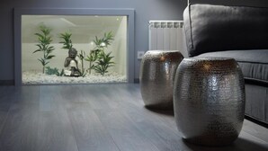 A modern living room with a dark wood floor, a grey sofa, and two textured vases on a coffee table. In the background, there's a framed picture of a Buddha statue surrounded by bamboo plants. - Lar de Donas (Meira)