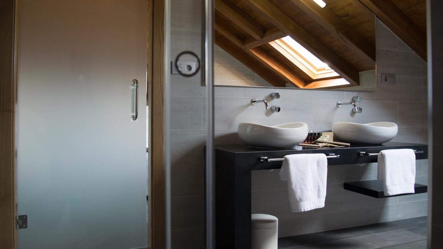A modern bathroom with two large white sinks, a skylight, and a wooden ceiling.