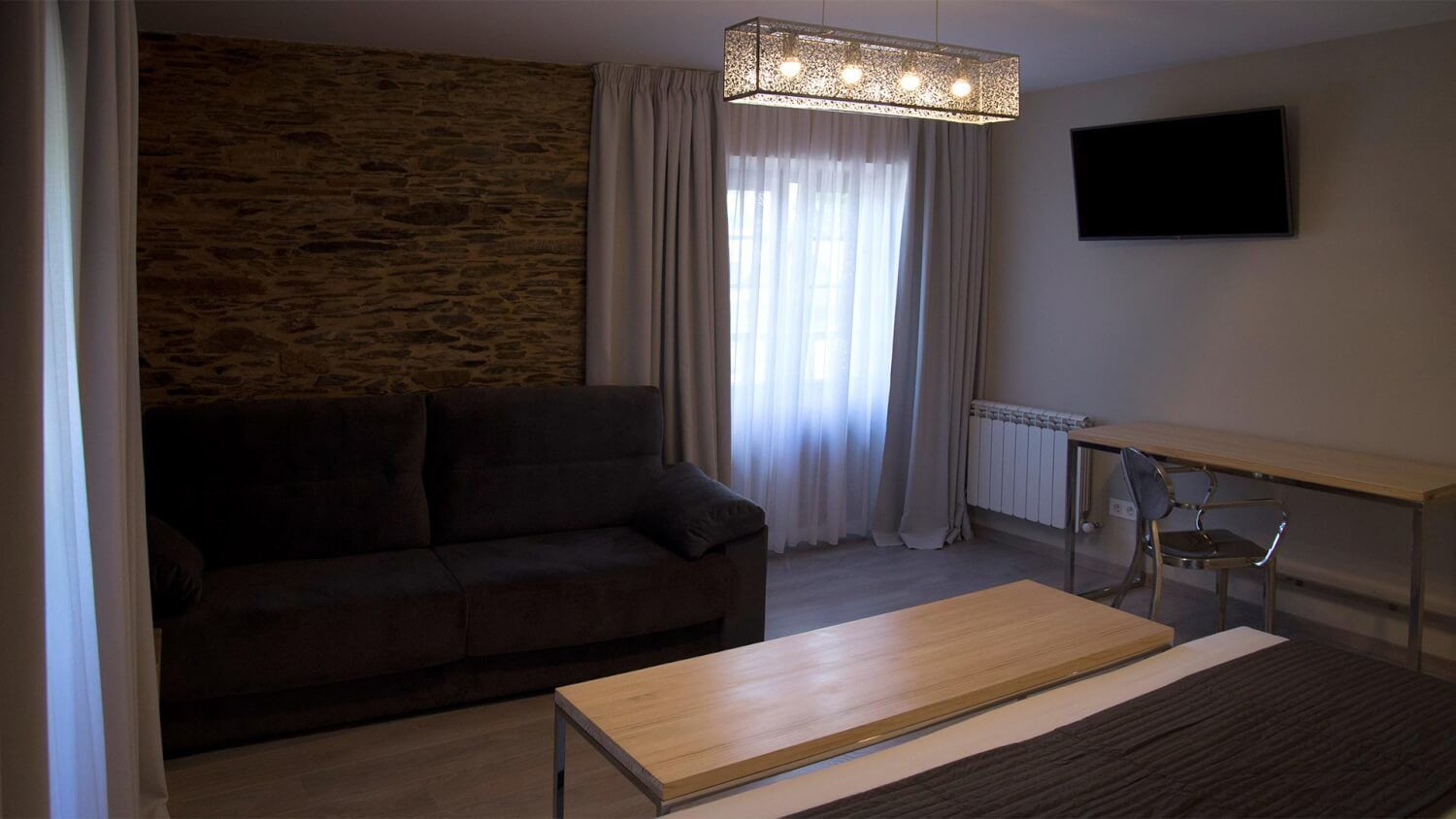 Una sala de estar con pared de piedra, un sofá oscuro, una mesa de centro de madera, una mesa de vidrio con una silla y un televisor montado en la pared.