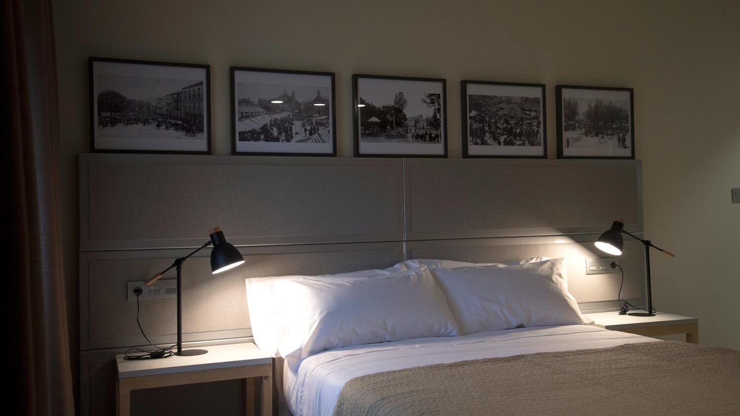 Habitación de hotel con una cama, dos lámparas de noche, un cabecero y cuatro fotografías en blanco y negro enmarcadas en la pared.