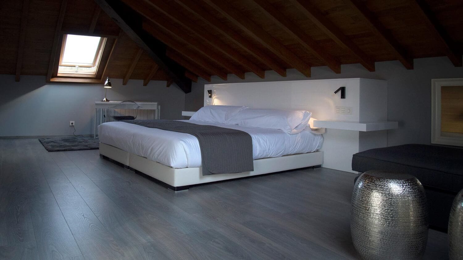 Un dormitorio con una cama, una mesita de noche, una lámpara, un jarrón grande y un tragaluz.