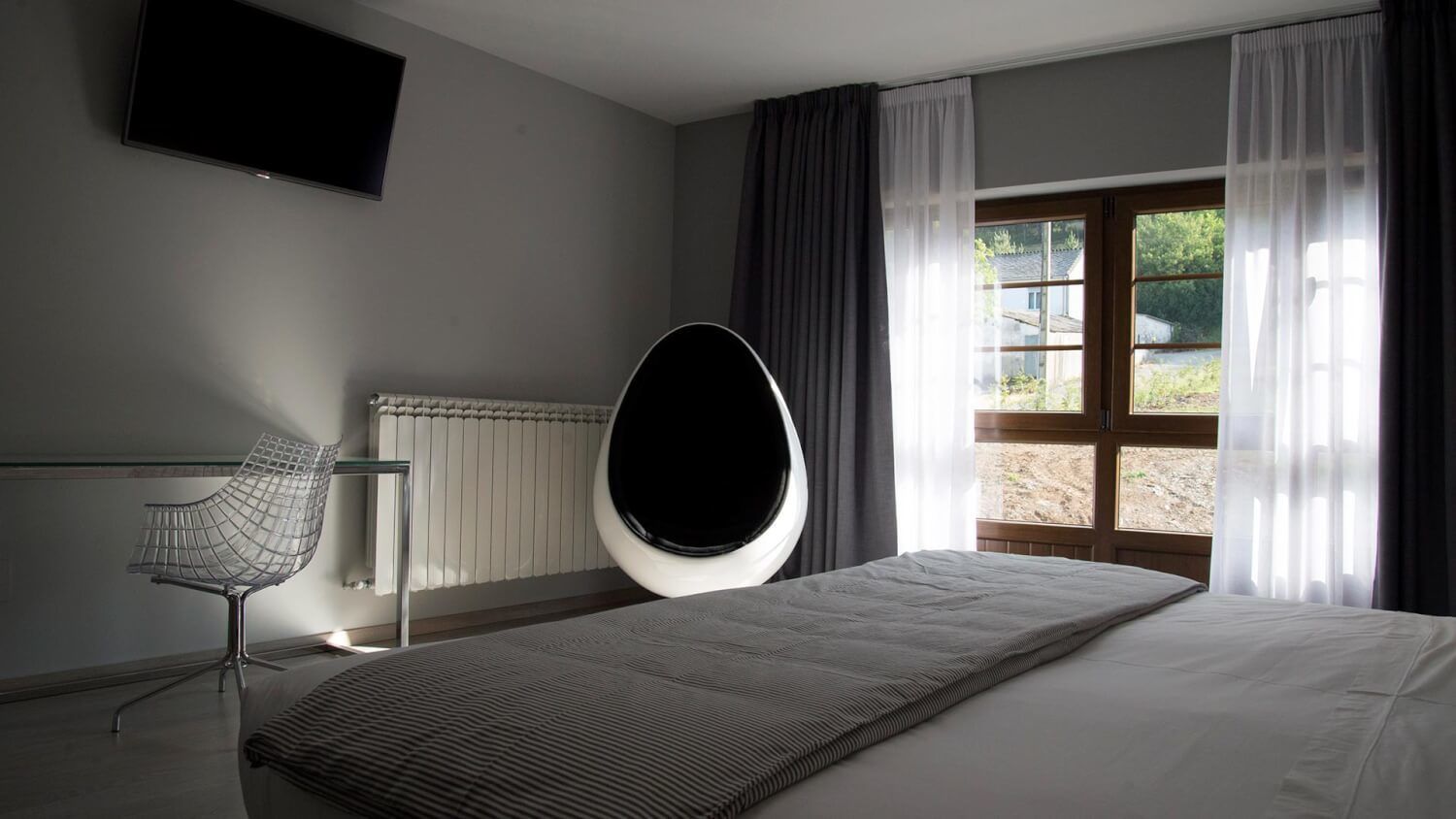 Una camera da letto con un letto, una sedia, un termosifone, una finestra con tende e una televisione appesa al muro.