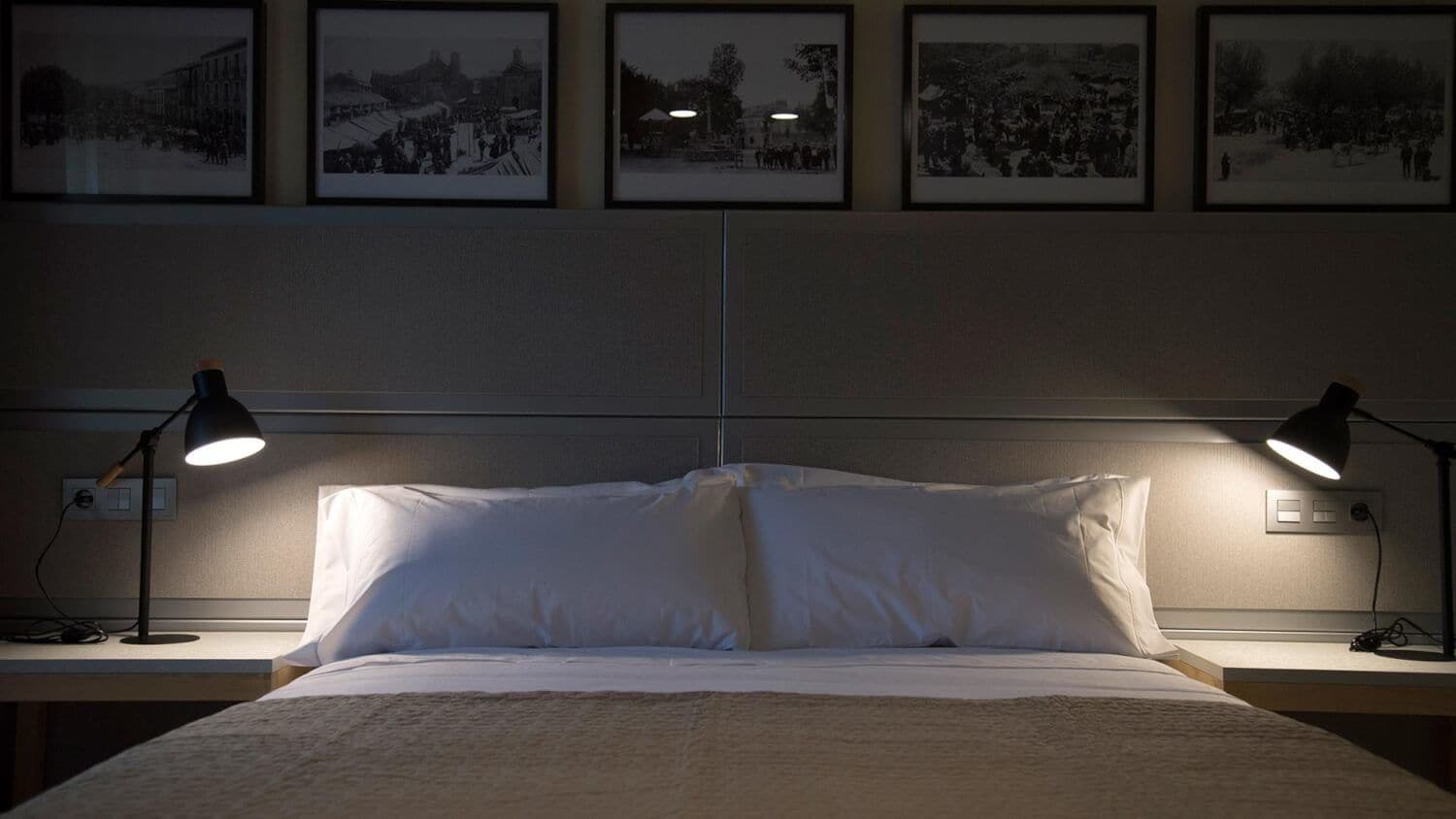 A neatly made bed with white linens, flanked by two bedside lamps on a nightstand. Above the bed, there are four framed black-and-white photographs on the wall.