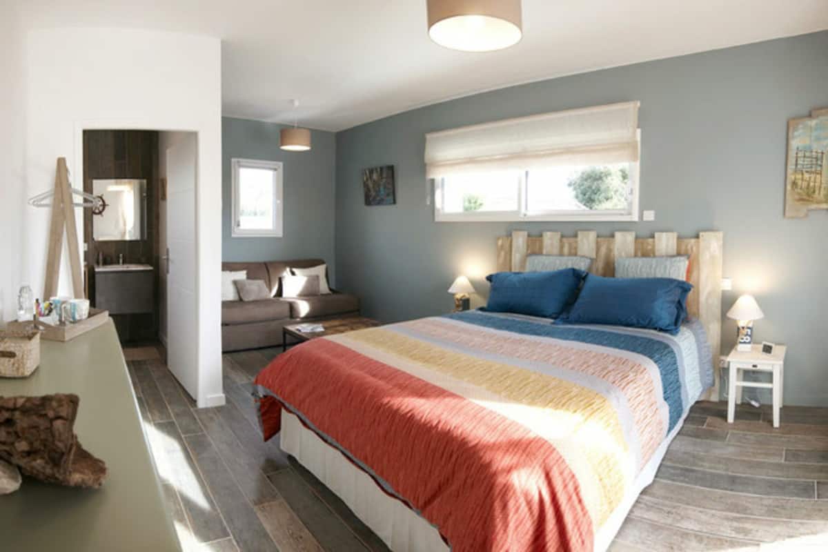 Een slaapkamer met een groot bed, een nachtkastje, een raam met jaloezieën, een spiegel en een deur naar een andere kamer.