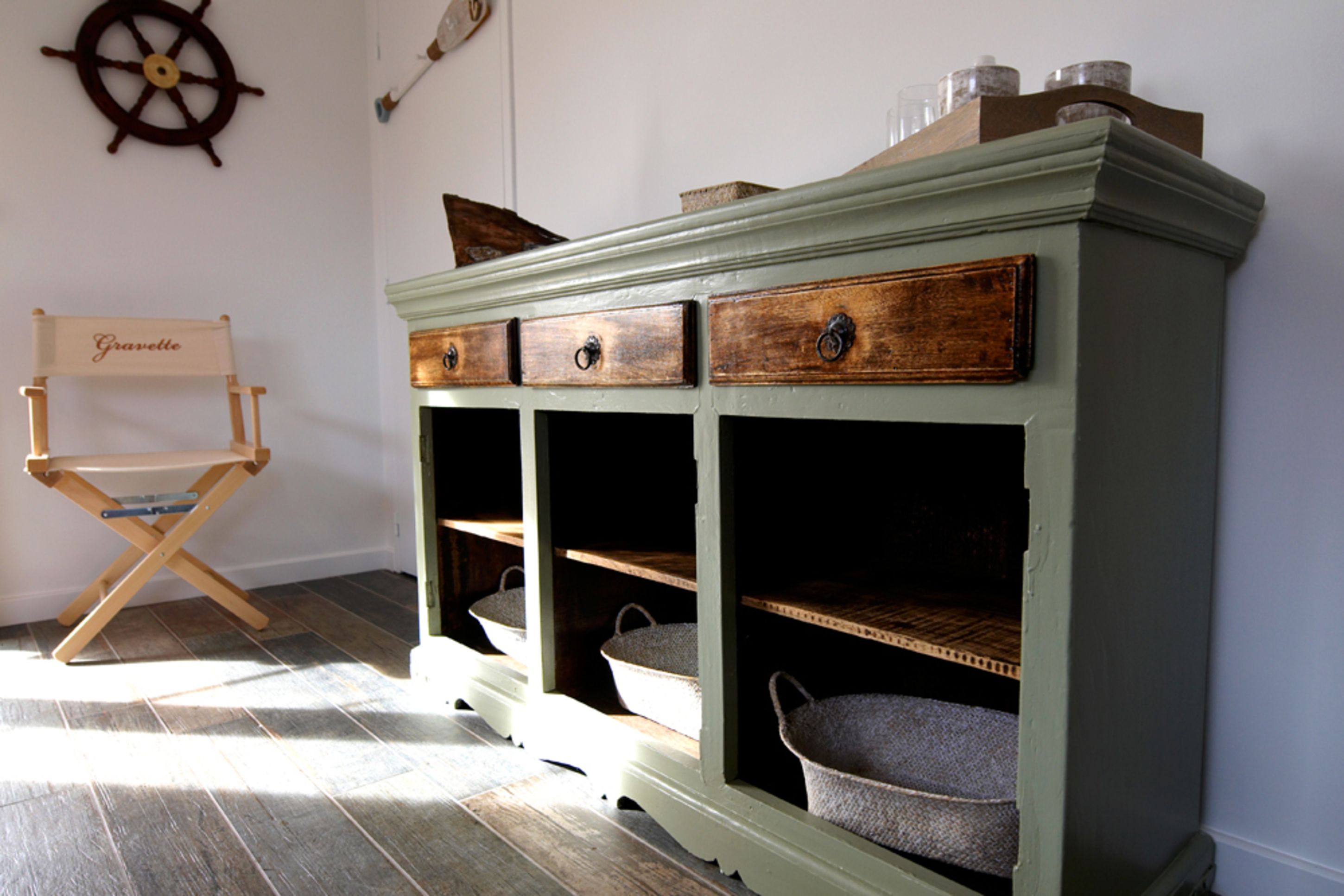 Un mobile verde con tre cassetti e due ripiani, una sedia di legno con un cartello e una ruota di timone appesa al muro.