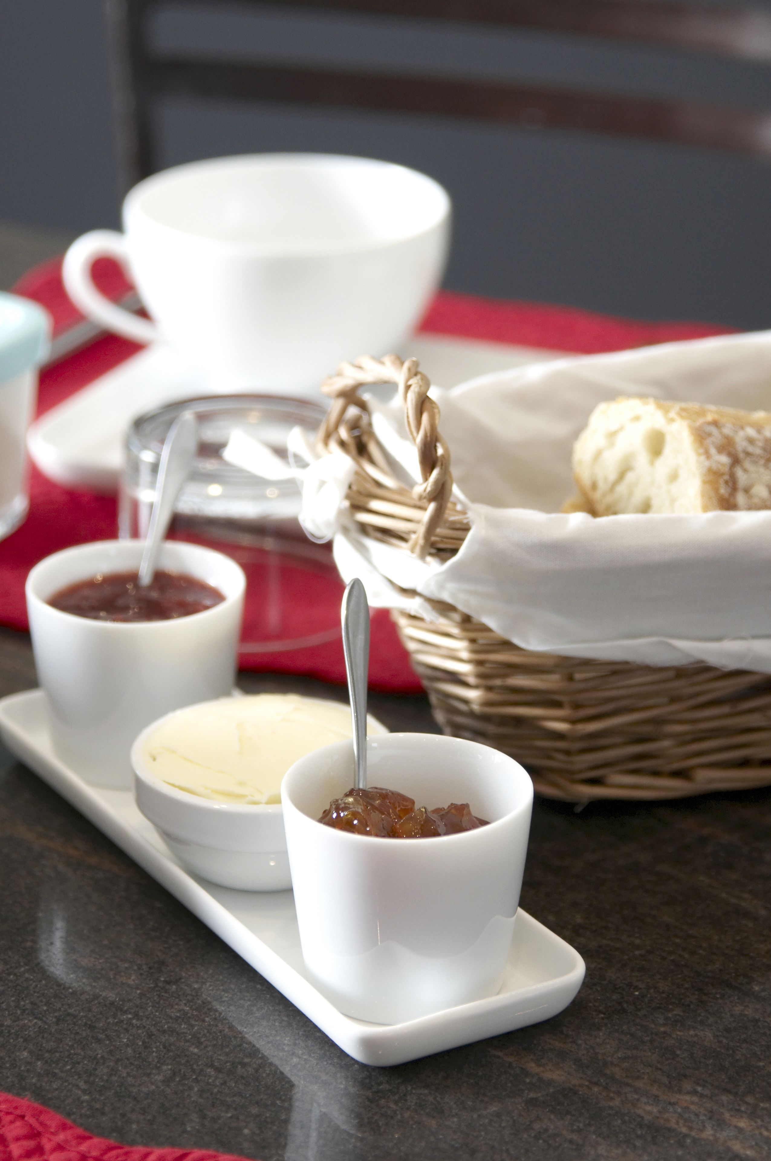 A safata amb tres tasses blanques que contenen diferents melmelades, un cistell de pa i un full vermell sobre una superfície fosca.