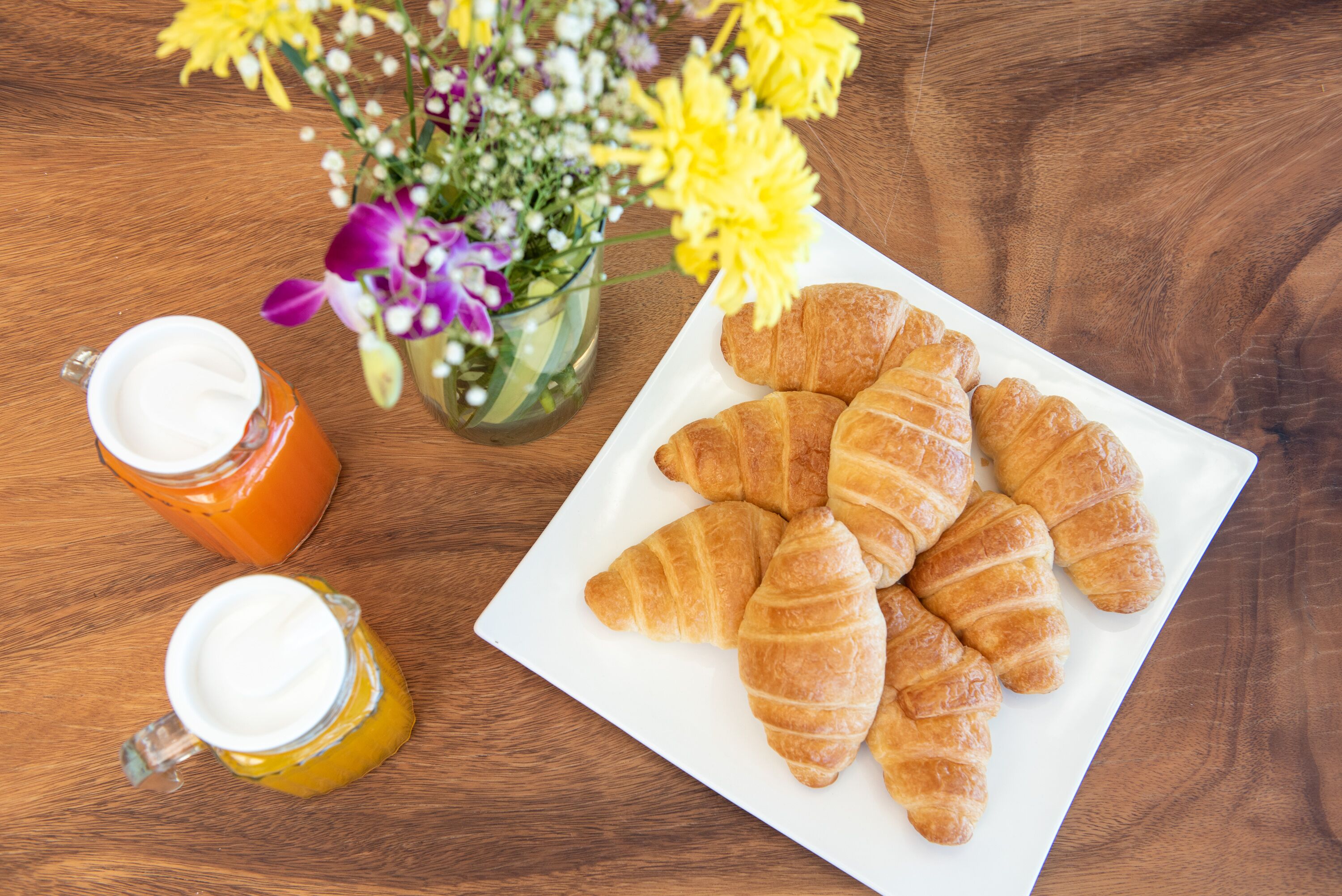 Ett träbord med en vit tallrik full av croissanter, två glas med en vit vätska och en vas med gula och lila blommor.