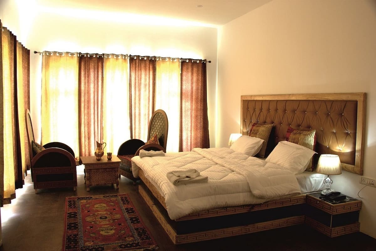 A hotel room with a large bed, a bedside table with a lamp, a chair, a small table with a vase, and a decorative rug.