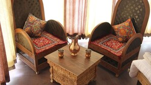 Dos sofás de madera ornamentados con tallas intrincadas y cojines coloridos, una mesa de centro de madera con una jarrón y tazas, y una cortina estampada al fondo.