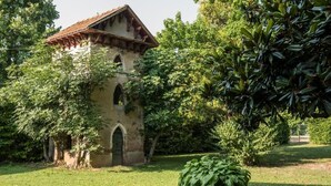 A historic building with a tiled roof, arched windows, and a small door, surrounded by lush greenery and a well-maintained lawn. - Azienda Agricola Le Colombare (Nogarole Rocca)