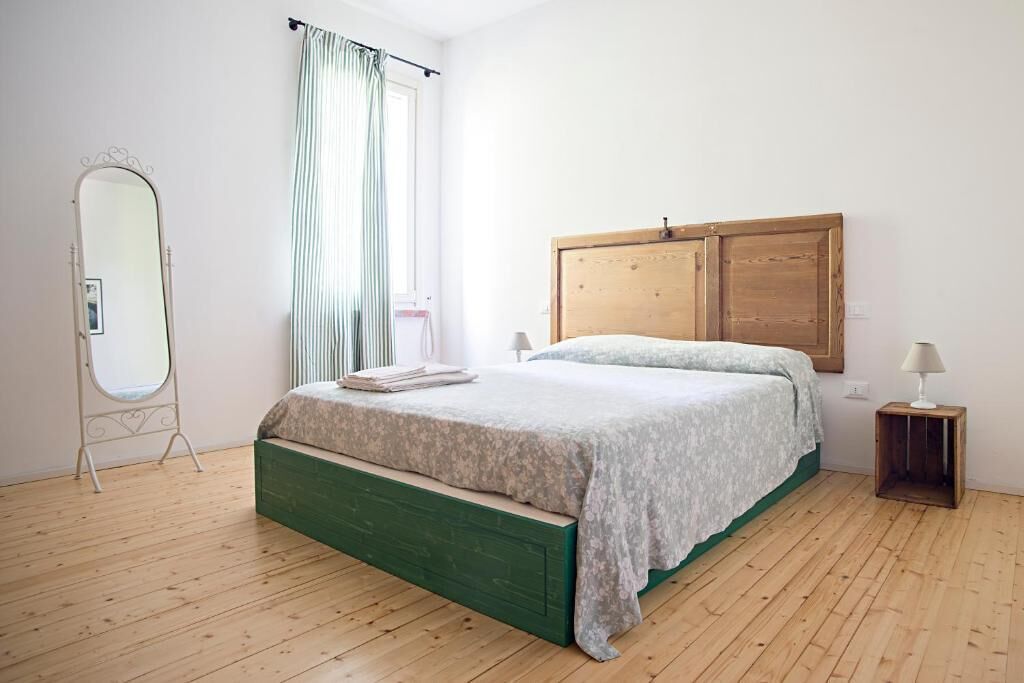 A bedroom with a wooden floor, a green bed frame, a wooden headboard, a bedside table with a lamp, and a full-length mirror.