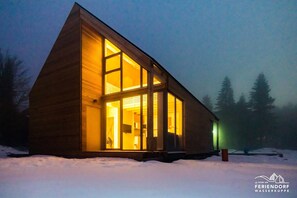 Una cabaña moderna de madera con amplios ventanales, ubicada en un paisaje nevado de noche.