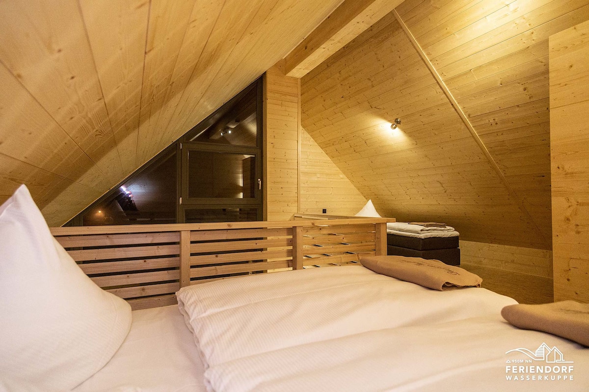 A cozy attic bedroom with a sloped ceiling, wooden walls, a bed with white bedding, and a small table with a lamp.