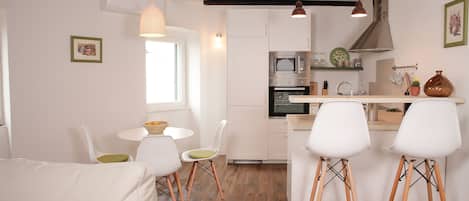 Una cocina moderna con armarios blancos, un microondas integrado y una campana extractora de acero inoxidable. Hay dos taburetes blancos y una mesa con sillas.