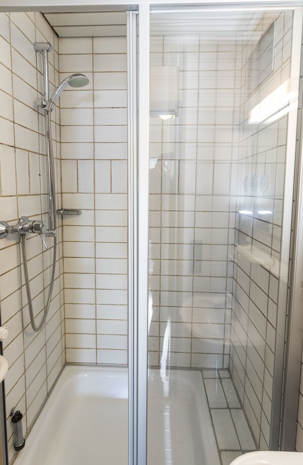 Un pequeño baño con revestimiento de azulejos, puerta de vidrio, ducha de mano y grifo empotrado.