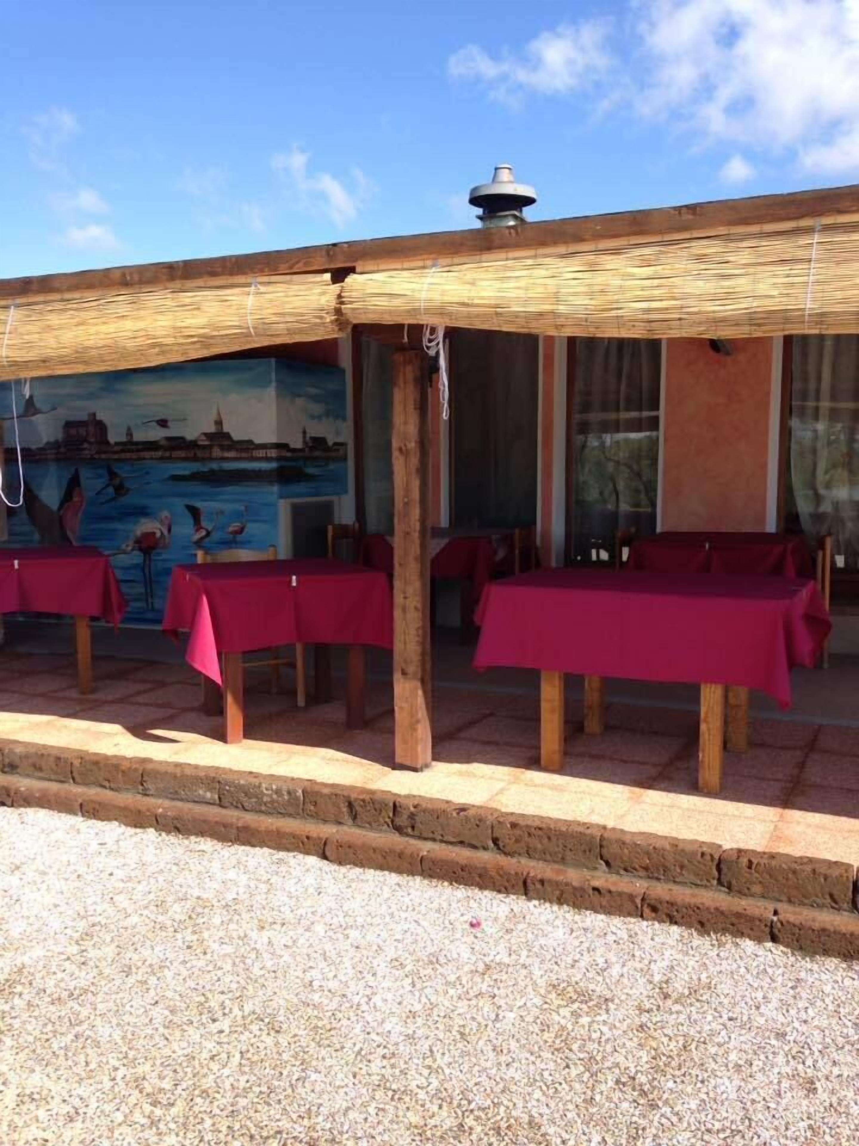 An outdoor seating area with tables covered in red tablecloths, wooden pillars, and a mural of a coastal scene.