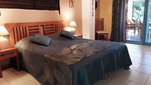 Een slaapkamer met een houten hoofdbord, een bed met een donkerblauwe deken, twee nachtlampjes en uitzicht buiten door een glazen deur.
