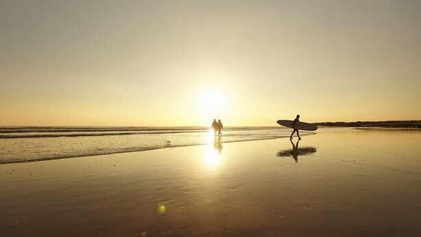 Eine Strandlandschaft bei Sonnenuntergang, zwei Personen gehen spazieren und eine Person trägt einen Surfbrett.