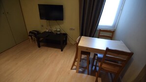 Habitació petita amb una taula i cadires de fusta, una televisió penjada a la paret i una finestra amb cortines.