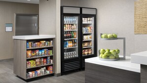 A hotel lobby with a reception desk, a refrigerator stocked with beverages and snacks, and a display of green apples. - Home2 Suites by Hilton Phoenix Airport South (Phoenix)
