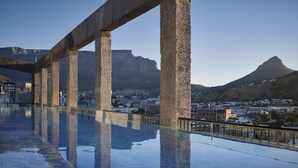 An infinity pool with clear water reflecting the surrounding architecture, a view of a cityscape, and a prominent mountain in the background. - The Silo Hotel (Cape Town)