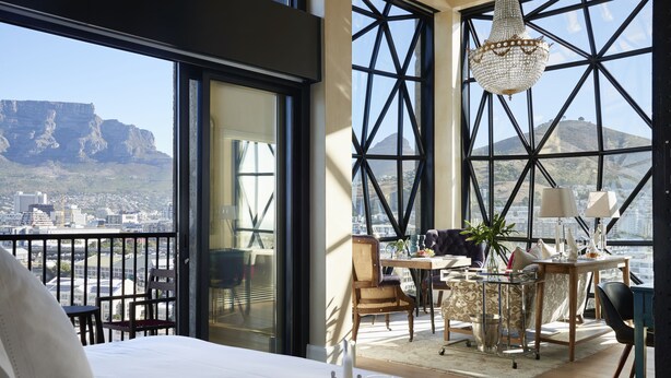 A hotel room with a large window offering a city view, a dining area with a table and chairs, and a bed with a tray of tea. - The Silo Hotel (Cape Town)