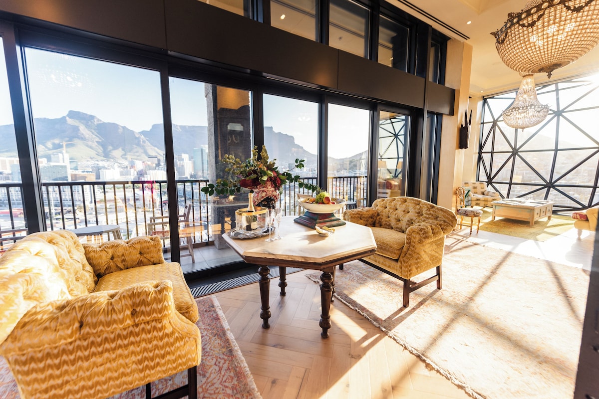 A spacious balcony with a view of a cityscape and mountains, furnished with a sofa, armchairs, and a table set for a meal.