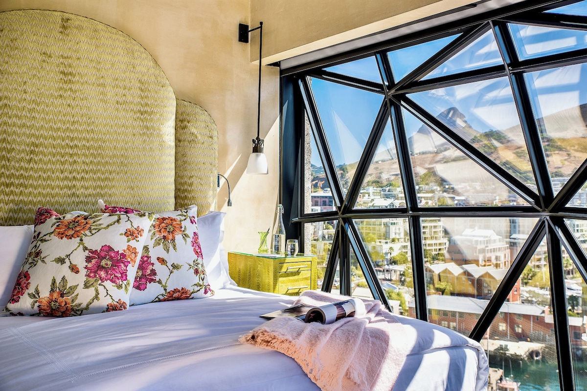 A bed with floral pillows, a bedside table with a bottle and glasses, and a large window with a view of buildings.